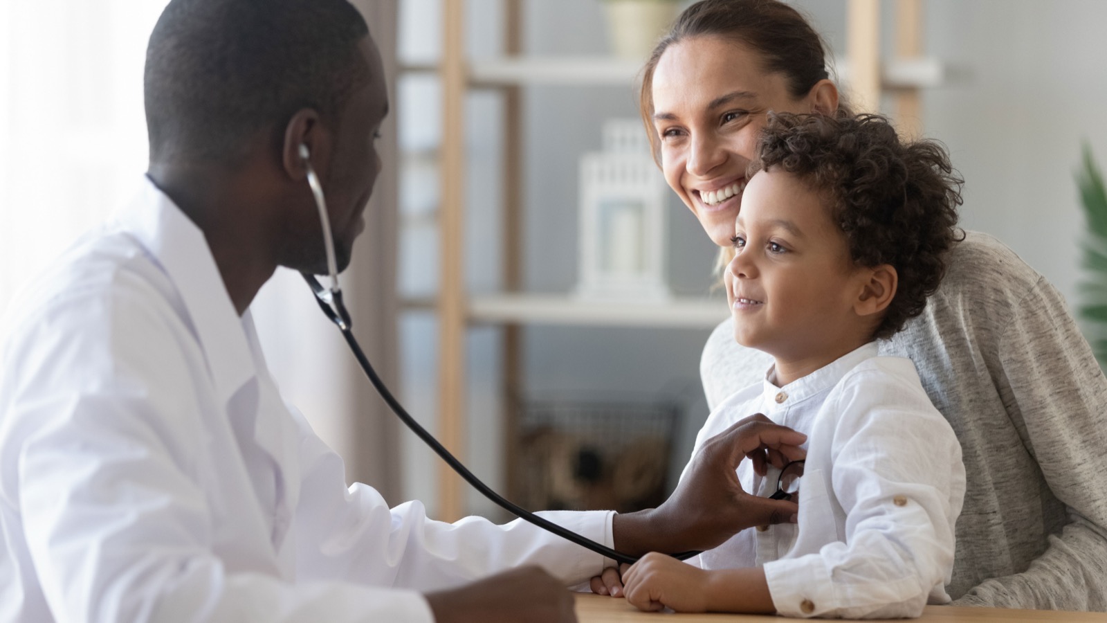 A doctor listening to a young child's chest while the child sits with a parent
